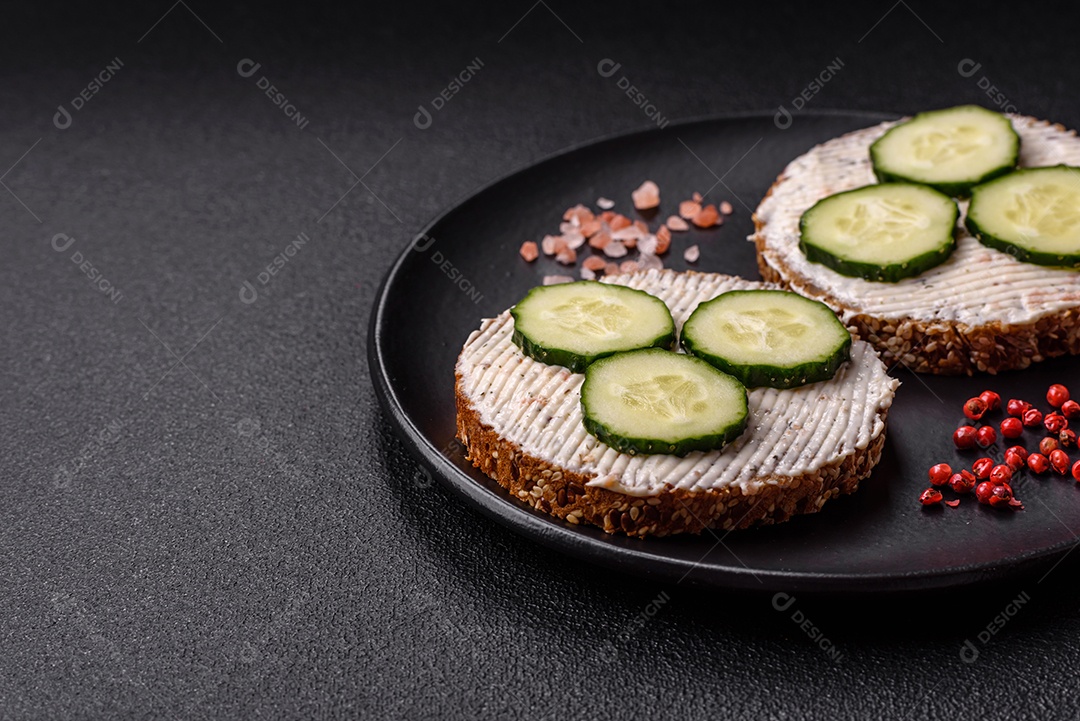 Delicioso sanduíche vegetariano com torradas grelhadas, cream cheese, pepinos e sementes e grãos sobre um fundo escuro de concreto