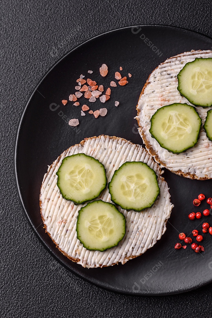 Delicioso sanduíche vegetariano com torradas grelhadas, cream cheese, pepinos e sementes e grãos sobre um fundo escuro de concreto