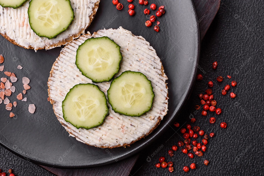 Delicioso sanduíche vegetariano com torradas grelhadas, cream cheese, pepinos e sementes e grãos sobre um fundo escuro de concreto