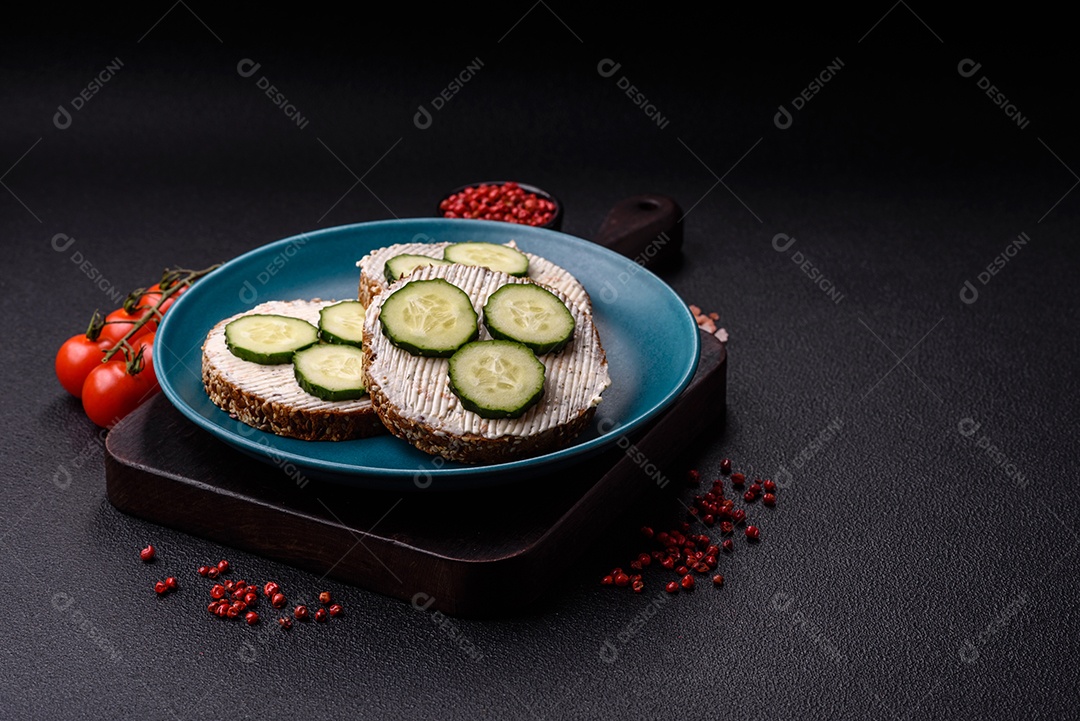 Delicioso sanduíche vegetariano com torradas grelhadas, cream cheese, pepinos e sementes e grãos sobre um fundo escuro de concreto
