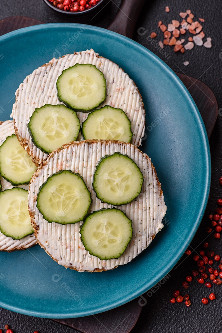 Delicioso sanduíche vegetariano com torradas grelhadas, cream cheese, pepinos e sementes e grãos sobre um fundo escuro de concreto