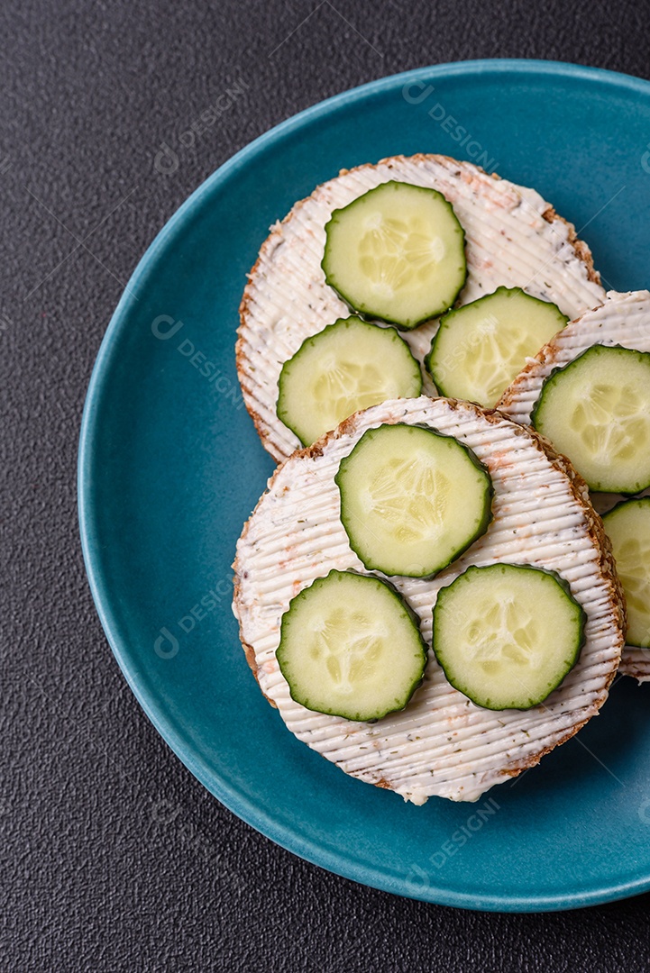Delicioso sanduíche vegetariano com torradas grelhadas, cream cheese, pepinos e sementes e grãos sobre um fundo escuro de concreto