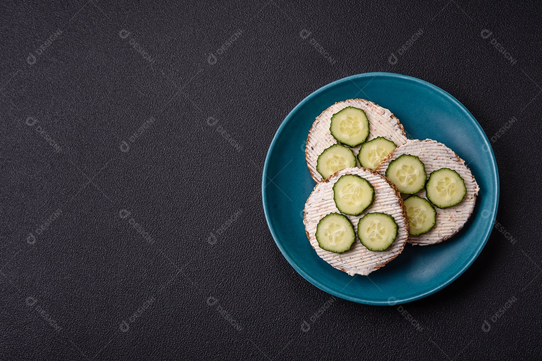 Delicioso sanduíche vegetariano com torradas grelhadas, cream cheese, pepinos e sementes e grãos sobre um fundo escuro de concreto