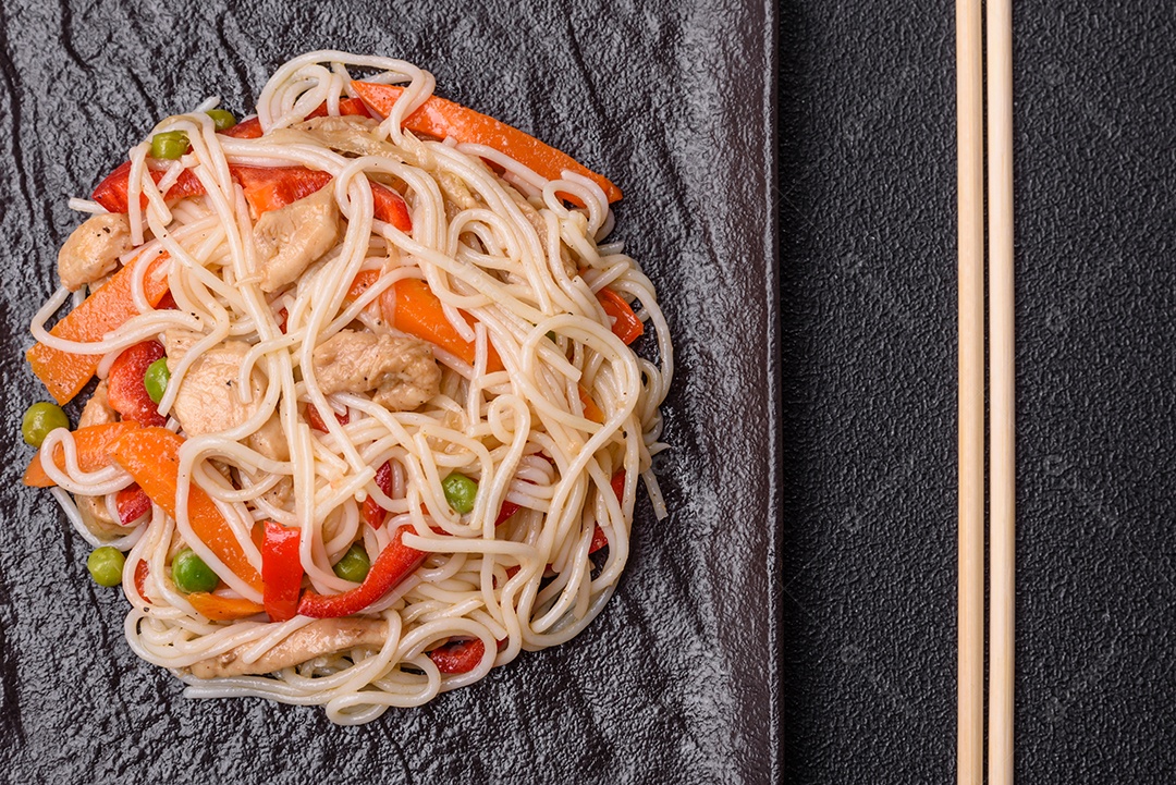 Delicioso macarrão de arroz ou udon com frango, cenoura, pimenta, sal, especiarias e ervas sobre fundo escuro de concreto