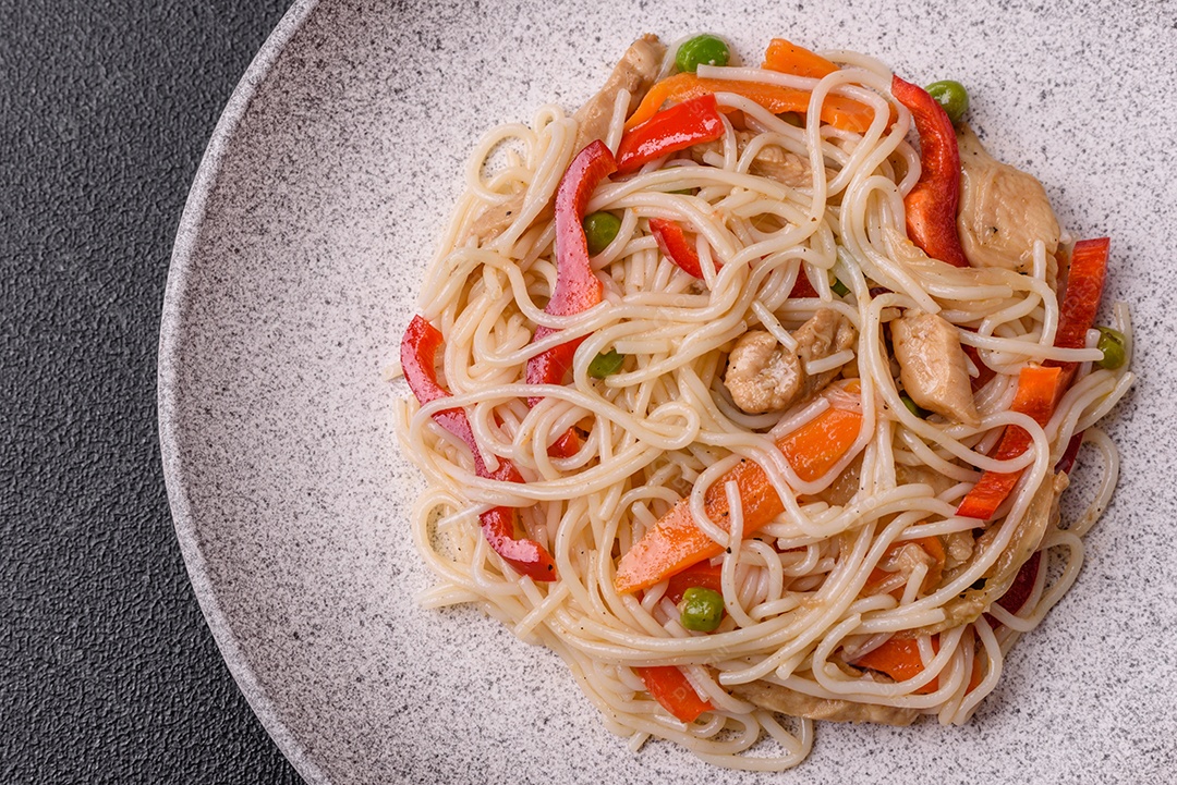 Delicioso macarrão de arroz ou udon com frango, cenoura, pimenta, sal, especiarias e ervas sobre fundo escuro de concreto