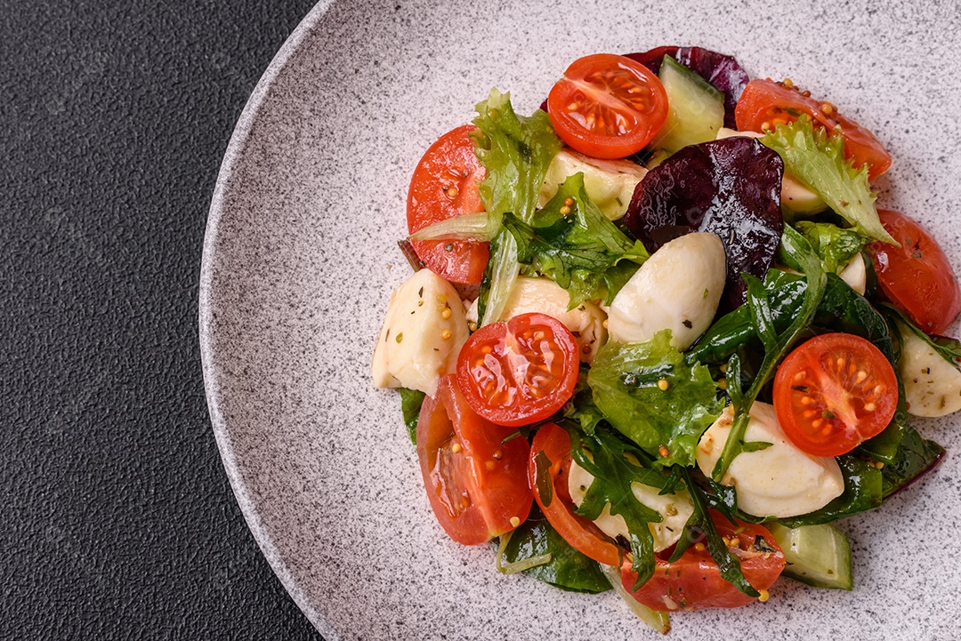 Deliciosa salada caprese fresca com mussarela, tomate, verduras com sal, especiarias e ervas sobre fundo escuro de concreto