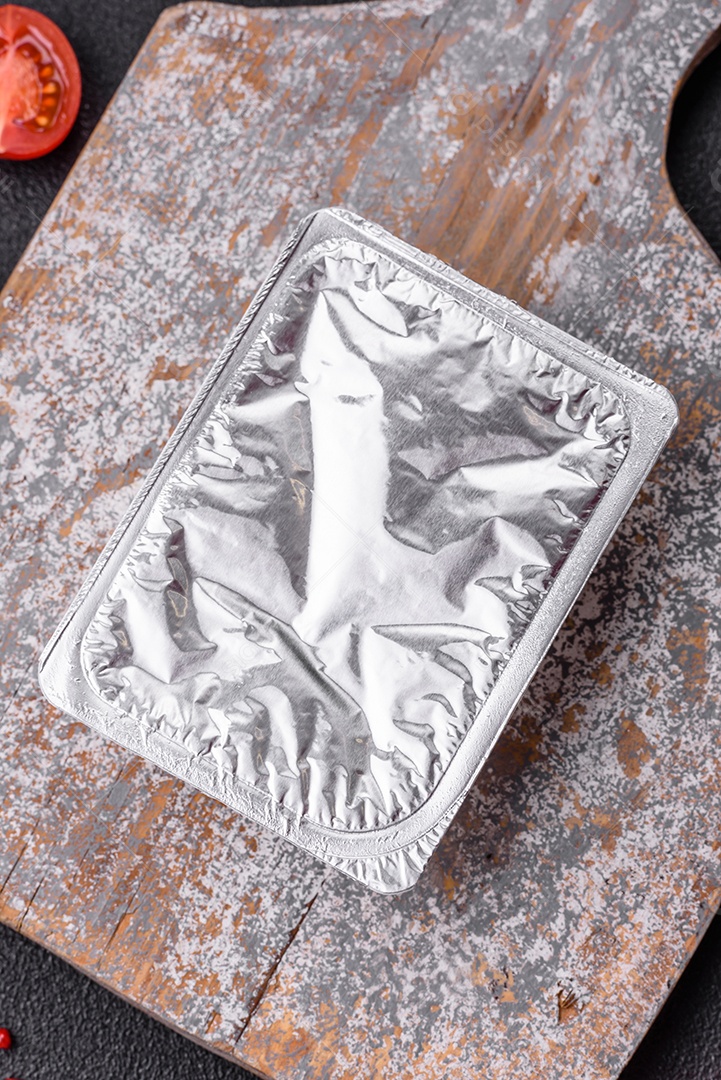 Caixa plástica retangular com iogurte ou queijo hermeticamente selada com tampa de alumínio sobre fundo escuro de concreto