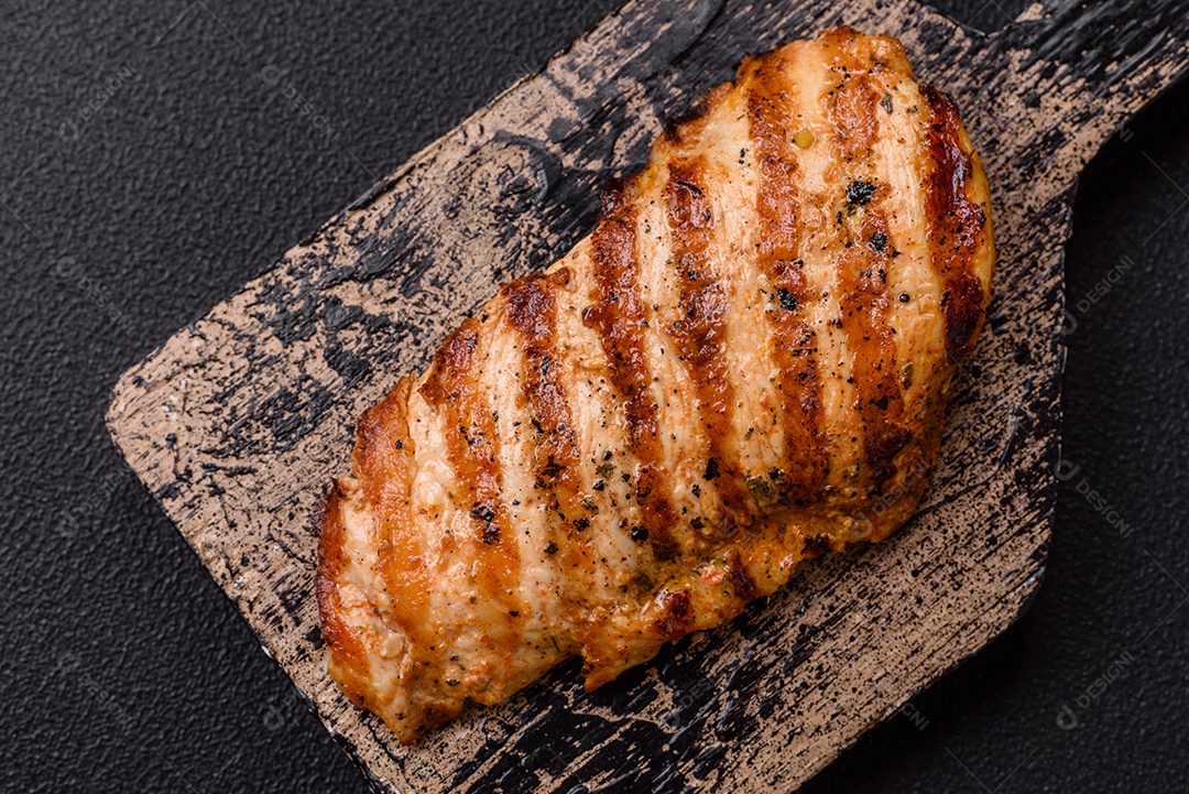 Delicioso filé de frango grelhado fresco com especiarias e ervas em um fundo escuro de concreto. Um prato cozido no fogo