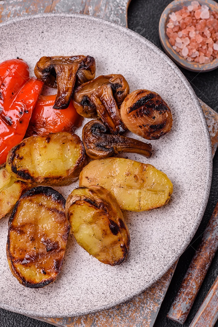 Deliciosos legumes grelhados, batatas, tomates, pimentões, berinjela com sal, especiarias e ervas em um fundo escuro de concreto