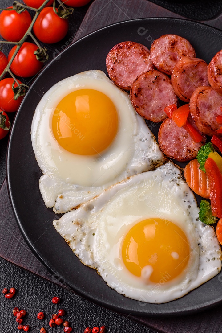 Delicioso café da manhã nutritivo com ovos fritos, salsicha, legumes cozidos no vapor com sal, especiarias e ervas sobre um fundo escuro de concreto