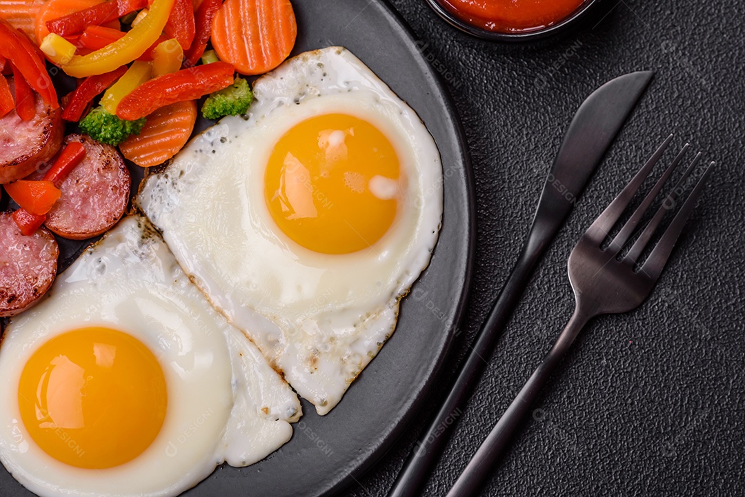 Delicioso café da manhã nutritivo com ovos fritos, salsicha, legumes cozidos no vapor com sal, especiarias e ervas sobre um fundo escuro de concreto