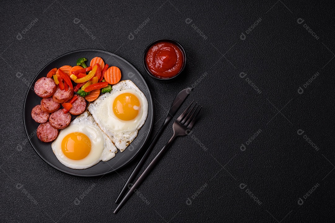Delicioso café da manhã nutritivo com ovos fritos, salsicha, legumes cozidos no vapor com sal, especiarias e ervas sobre um fundo escuro de concreto