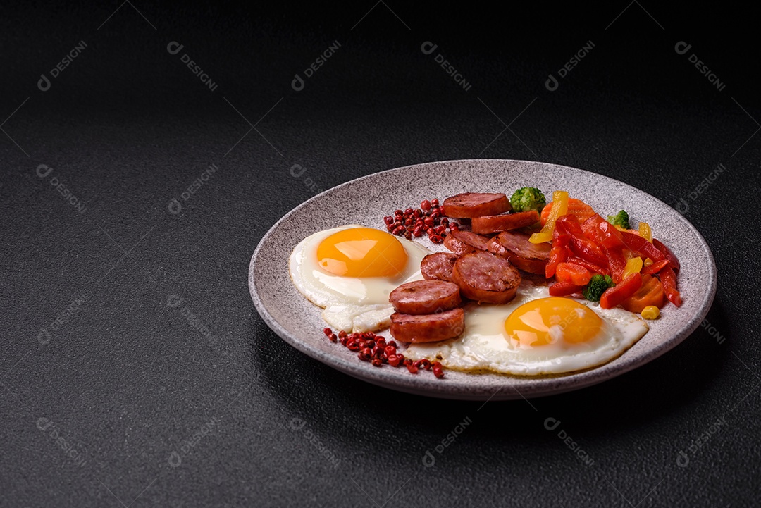 Delicioso café da manhã nutritivo com ovos fritos, salsicha, legumes cozidos no vapor com sal, especiarias e ervas sobre um fundo escuro de concreto