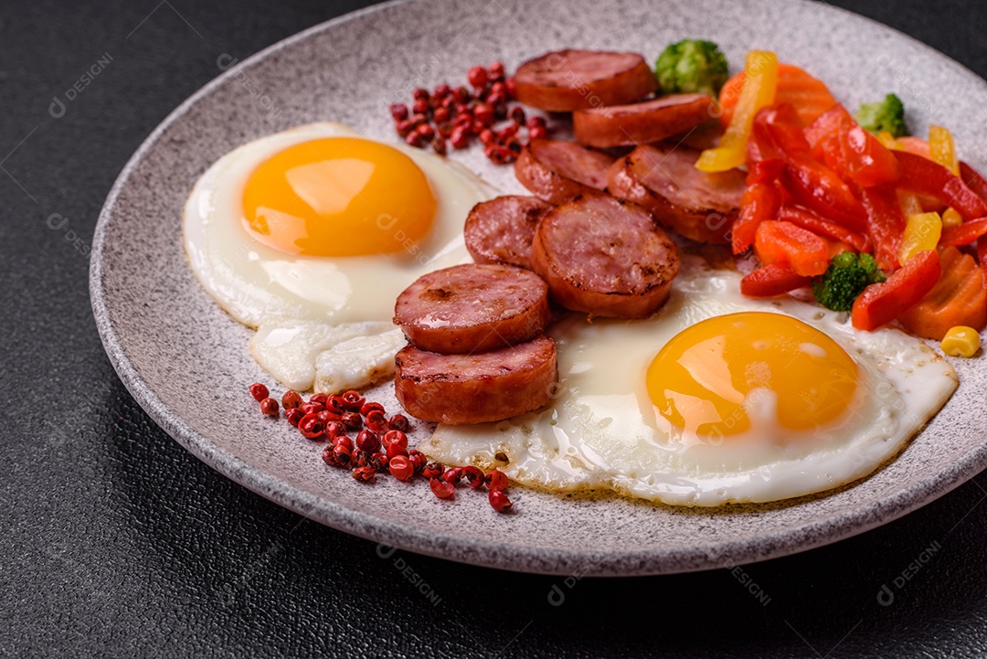 Delicioso café da manhã nutritivo com ovos fritos, salsicha, legumes cozidos no vapor com sal, especiarias e ervas sobre um fundo escuro de concreto