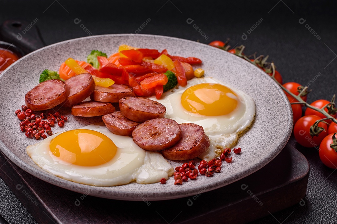 Delicioso café da manhã nutritivo com ovos fritos, salsicha, legumes cozidos no vapor com sal, especiarias e ervas sobre um fundo escuro de concreto