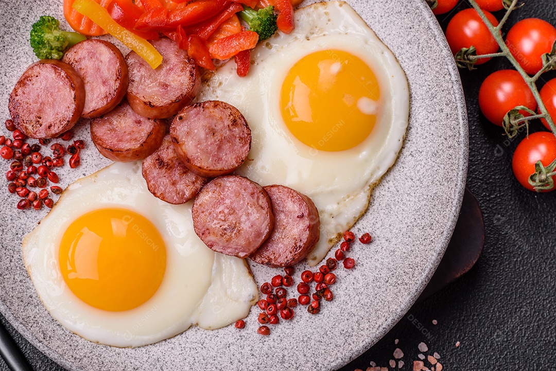 Delicioso café da manhã nutritivo com ovos fritos, salsicha, legumes cozidos no vapor com sal, especiarias e ervas sobre um fundo escuro de concreto