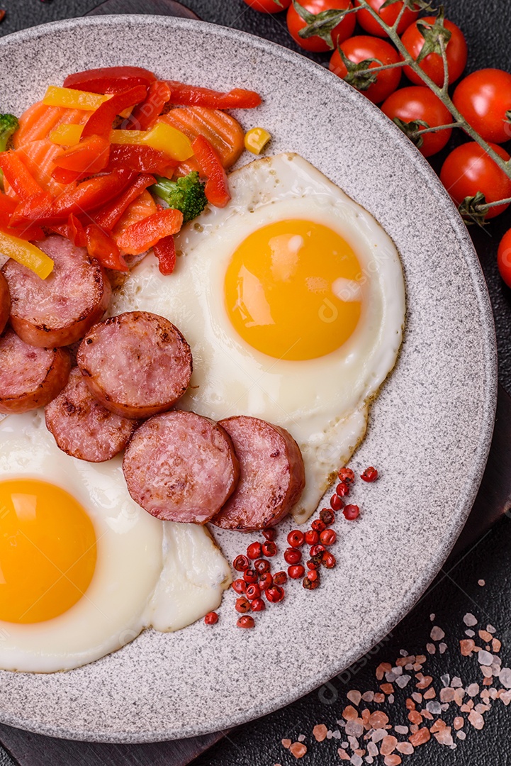 Delicioso café da manhã nutritivo com ovos fritos, salsicha, legumes cozidos no vapor com sal, especiarias e ervas sobre um fundo escuro de concreto