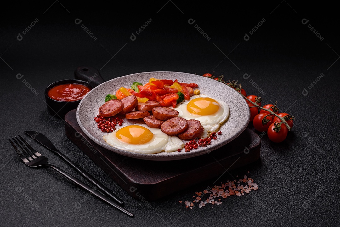 Delicioso café da manhã nutritivo com ovos fritos, salsicha, legumes cozidos no vapor com sal, especiarias e ervas sobre um fundo escuro de concreto
