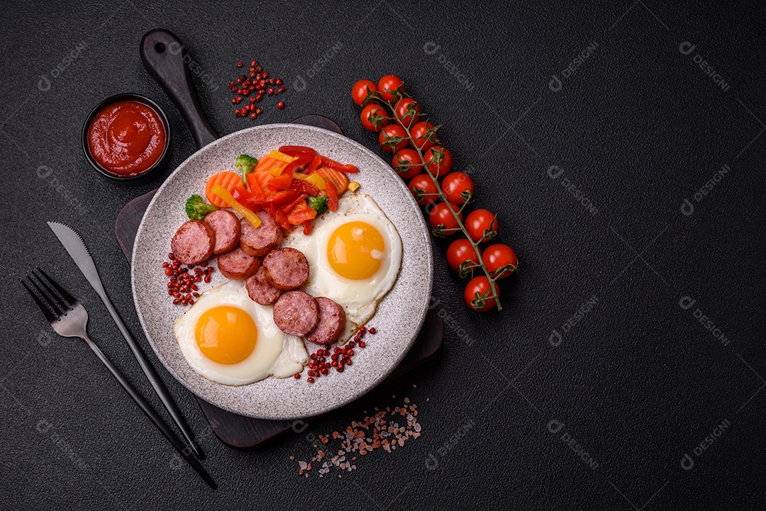 Delicioso café da manhã nutritivo com ovos fritos, salsicha, legumes cozidos no vapor com sal, especiarias e ervas sobre um fundo escuro de concreto