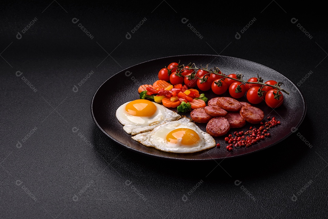 Delicioso café da manhã nutritivo com ovos fritos, salsicha, legumes cozidos no vapor com sal, especiarias e ervas sobre um fundo escuro de concreto