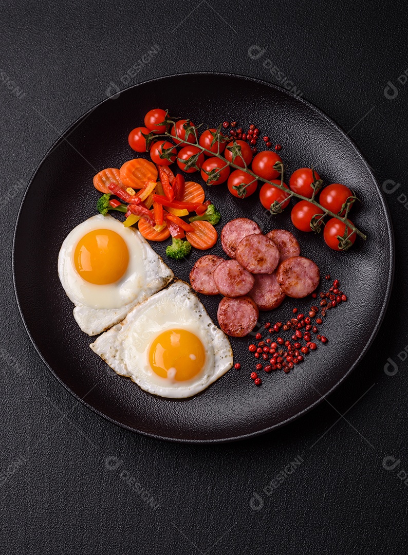 Delicioso café da manhã nutritivo com ovos fritos, salsicha, legumes cozidos no vapor com sal, especiarias e ervas sobre um fundo escuro de concreto