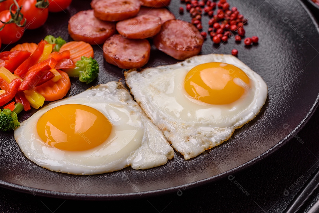 Delicioso café da manhã nutritivo com ovos fritos, salsicha, legumes cozidos no vapor com sal, especiarias e ervas sobre um fundo escuro de concreto