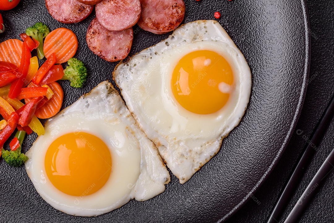 Delicioso café da manhã nutritivo com ovos fritos, salsicha, legumes cozidos no vapor com sal, especiarias e ervas sobre um fundo escuro de concreto