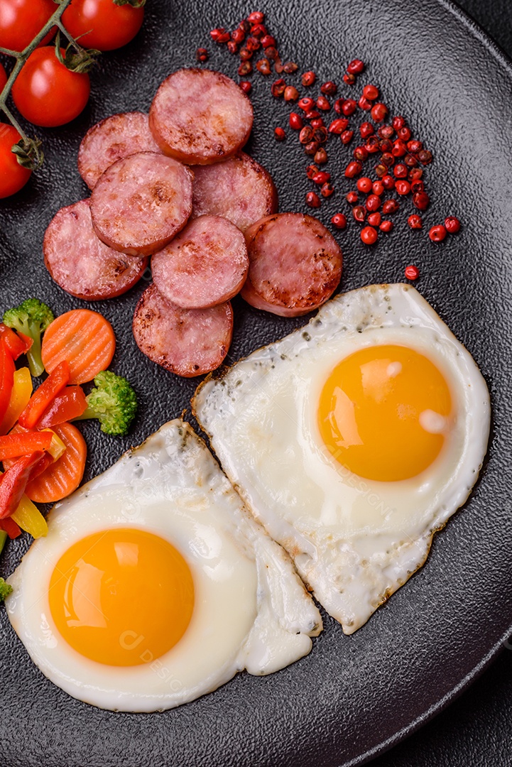 Delicioso café da manhã nutritivo com ovos fritos, salsicha, legumes cozidos no vapor com sal, especiarias e ervas sobre um fundo escuro de concreto