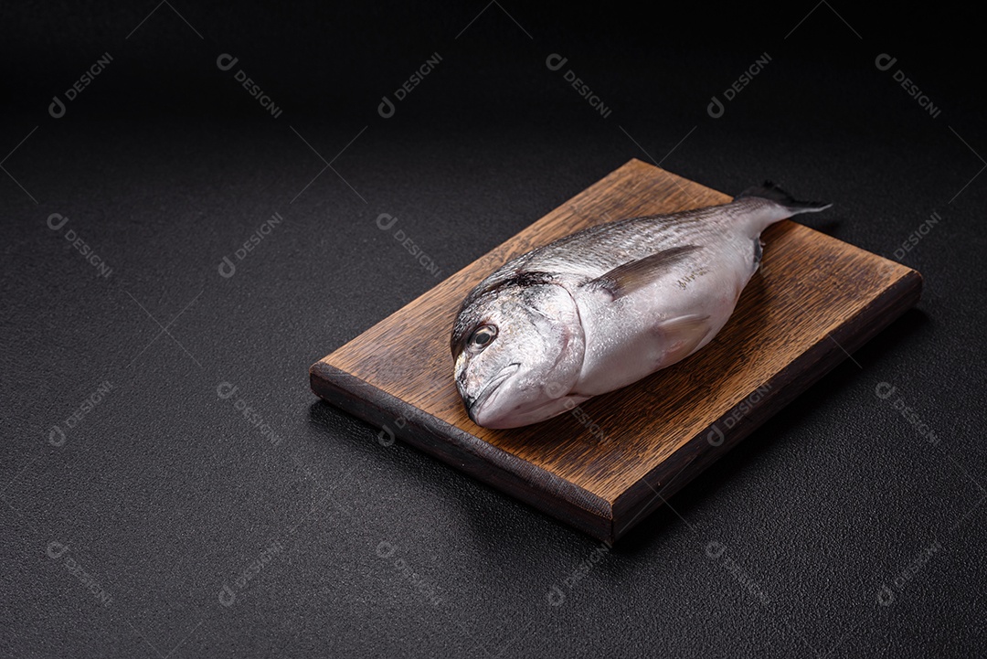 Peixe dourado cru inteiro com limão, sal, especiarias e ervas sobre um fundo escuro de concreto