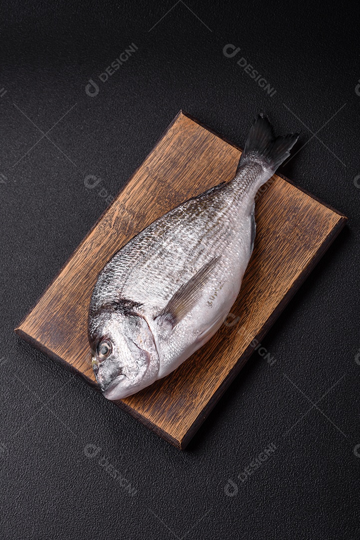Peixe dourado cru inteiro com limão, sal, especiarias e ervas sobre um fundo escuro de concreto