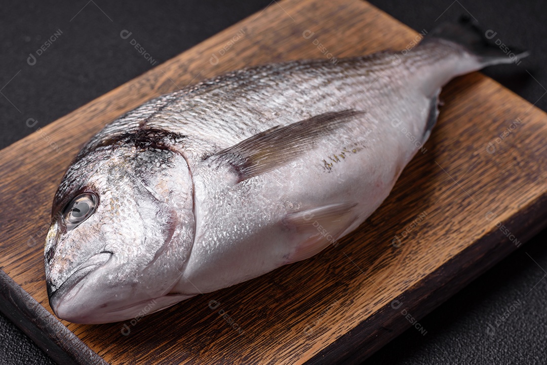 Peixe dourado cru inteiro com limão, sal, especiarias e ervas sobre um fundo escuro de concreto