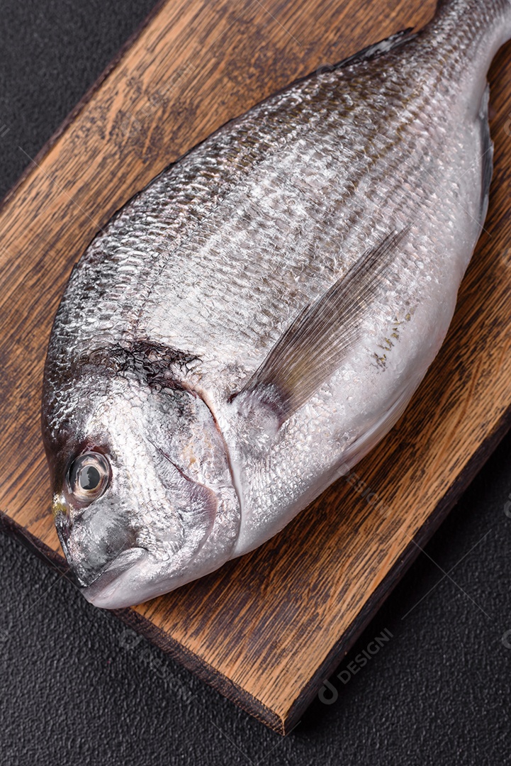 Peixe dourado cru inteiro com limão, sal, especiarias e ervas sobre um fundo escuro de concreto