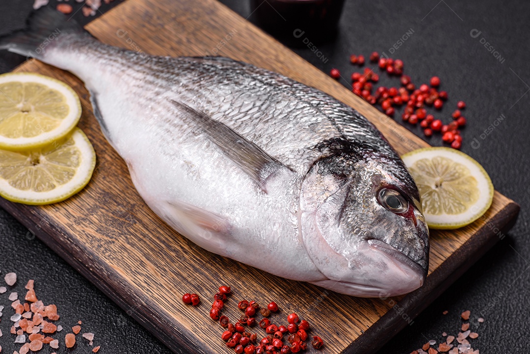 Peixe dourado cru inteiro com limão, sal, especiarias e ervas sobre um fundo escuro de concreto