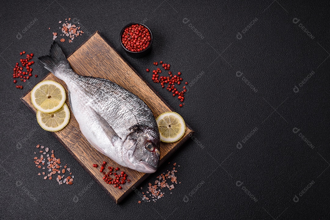 Peixe dourado cru inteiro com limão, sal, especiarias e ervas sobre um fundo escuro de concreto