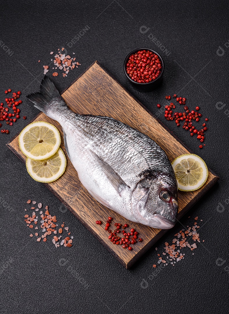 Peixe dourado cru inteiro com limão, sal, especiarias e ervas sobre um fundo escuro de concreto