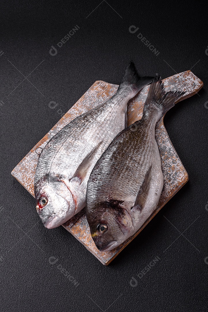 Peixe dourado cru inteiro com limão, sal, especiarias e ervas sobre um fundo escuro de concreto