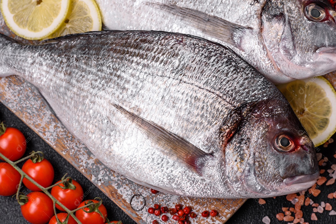 Peixe dourado cru inteiro com limão, sal, especiarias e ervas sobre um fundo escuro de concreto