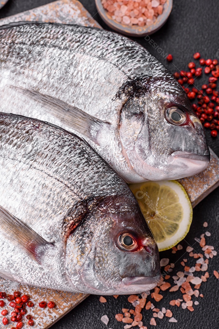 Peixe dourado cru inteiro com limão, sal, especiarias e ervas sobre um fundo escuro de concreto