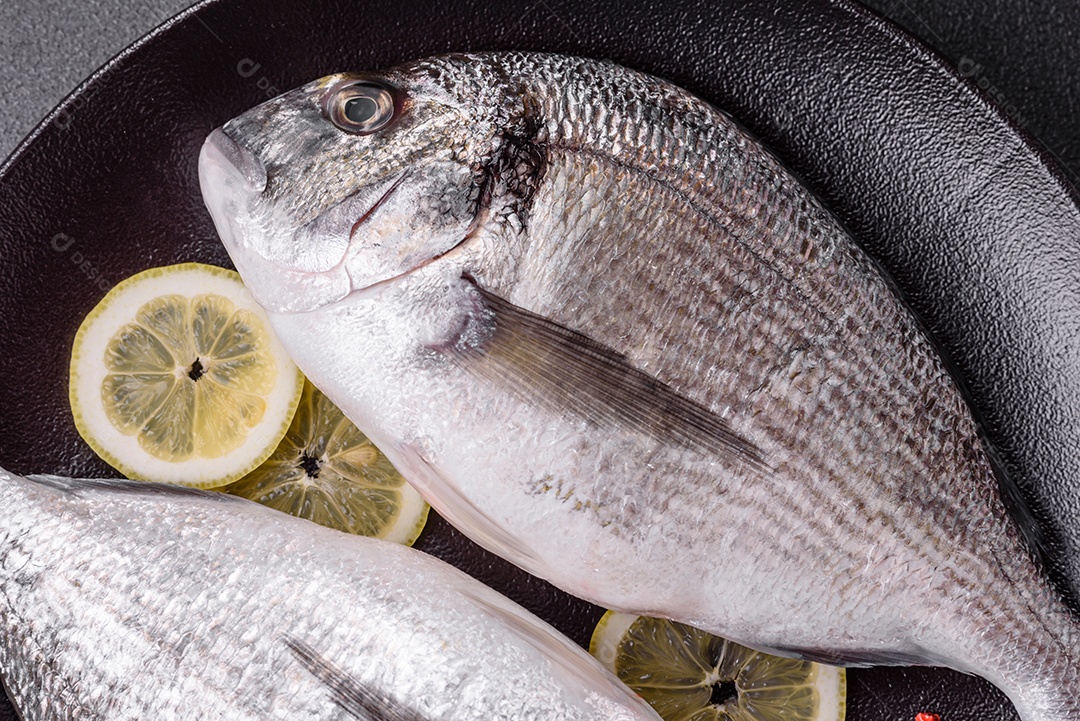 Peixe dourado cru inteiro com limão, sal, especiarias e ervas sobre um fundo escuro de concreto