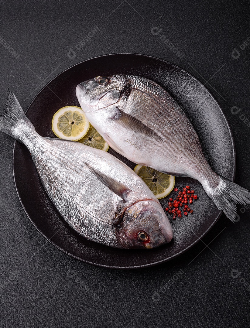 Peixe dourado cru inteiro com limão, sal, especiarias e ervas sobre um fundo escuro de concreto