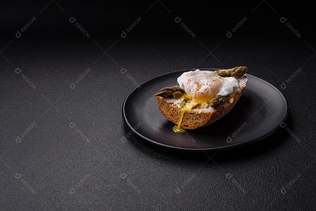 Delicioso café da manhã composto por ovos escalfados com torradas com cream cheese, aspargos, sal e especiarias em um fundo escuro de concreto