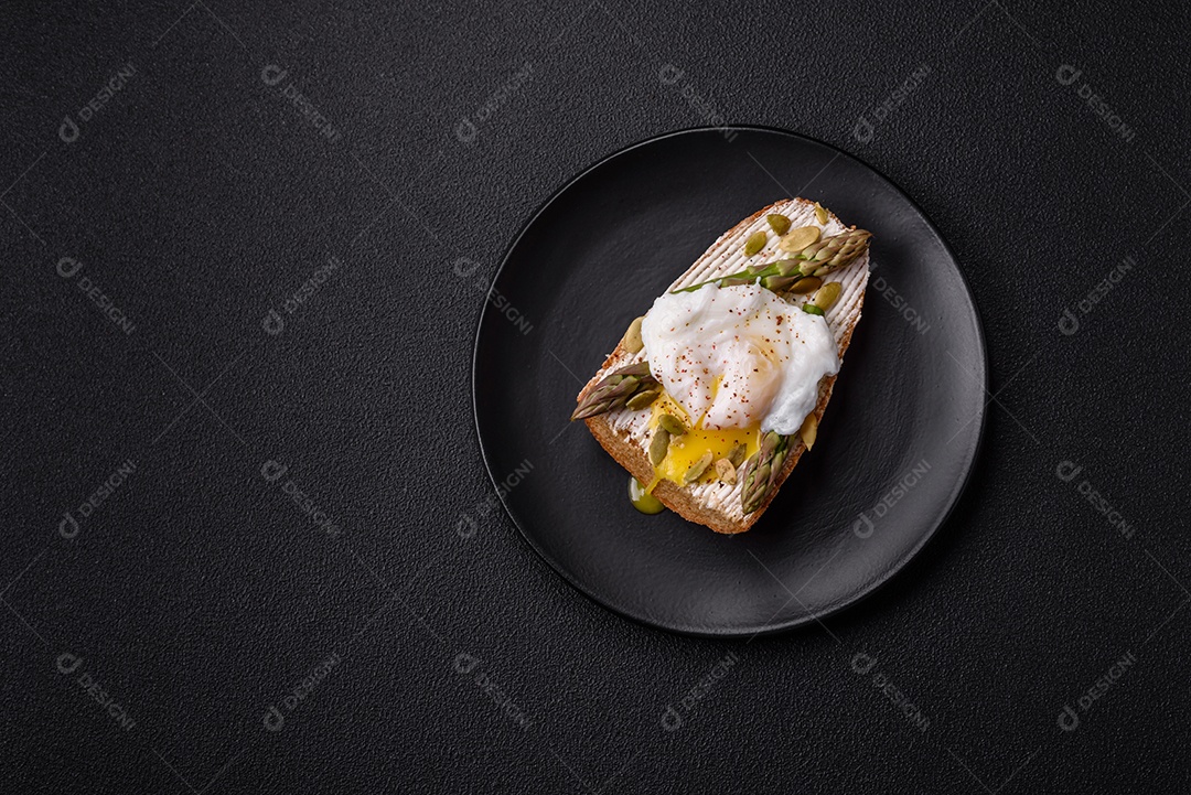 Delicioso café da manhã composto por ovos escalfados com torradas com cream cheese, aspargos, sal e especiarias em um fundo escuro de concreto