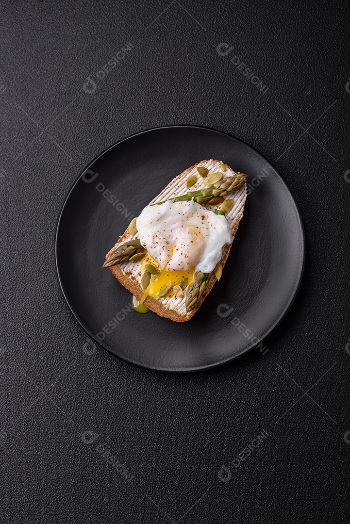 Delicioso café da manhã composto por ovos escalfados com torradas com cream cheese, aspargos, sal e especiarias em um fundo escuro de concreto