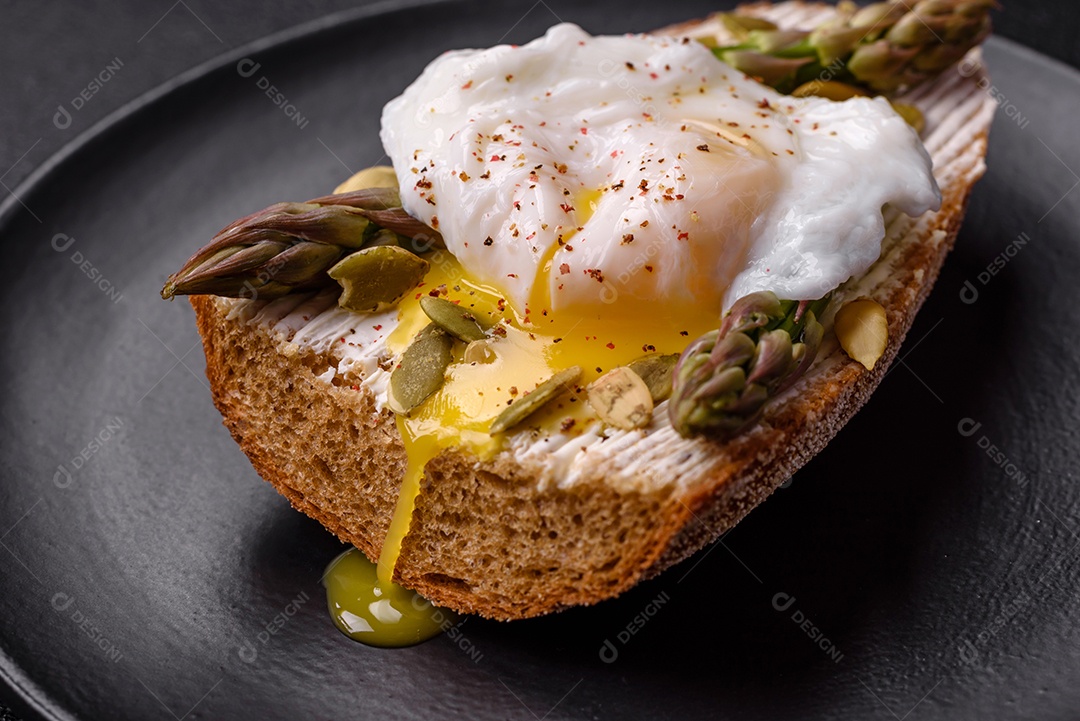 Delicioso café da manhã composto por ovos escalfados com torradas com cream cheese, aspargos, sal e especiarias em um fundo escuro de concreto