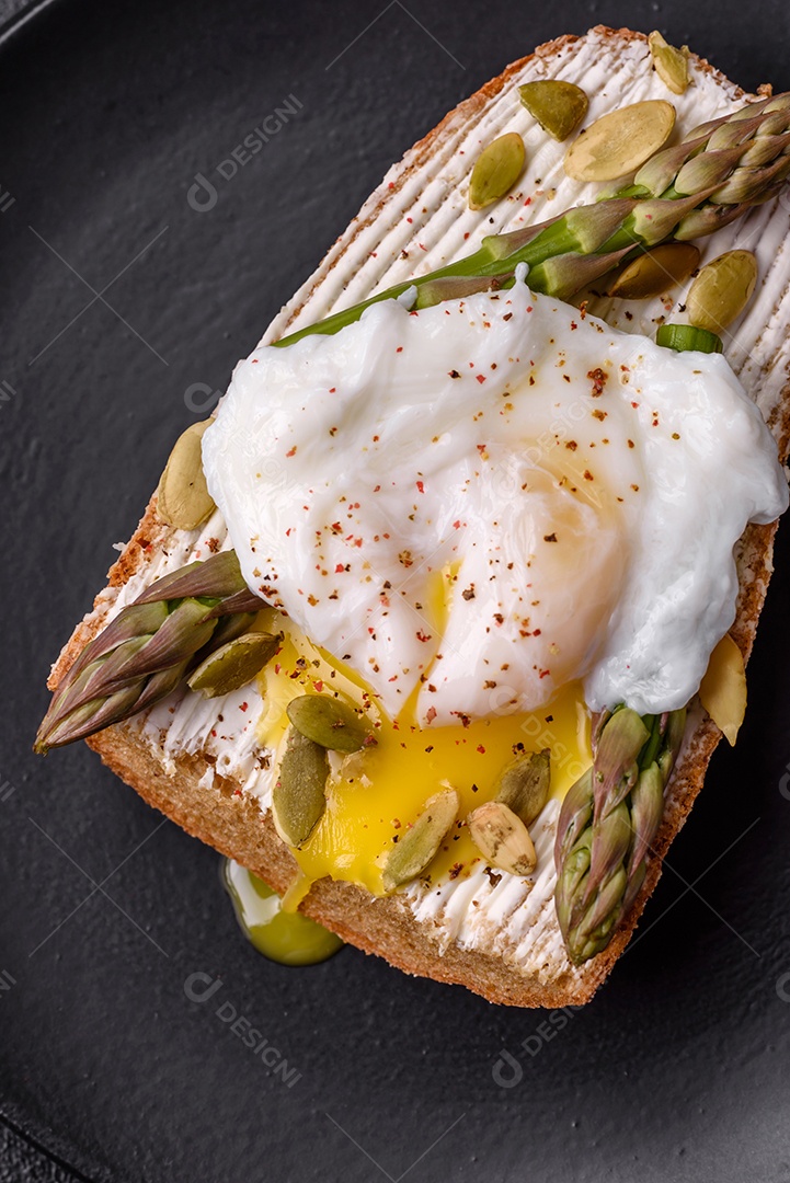 Delicioso café da manhã composto por ovos escalfados com torradas com cream cheese, aspargos, sal e especiarias em um fundo escuro de concreto