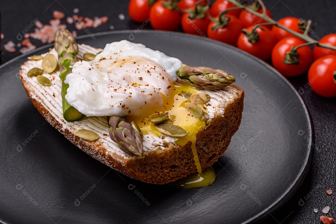 Delicioso café da manhã composto por ovos escalfados com torradas com cream cheese, aspargos, sal e especiarias em um fundo escuro de concreto