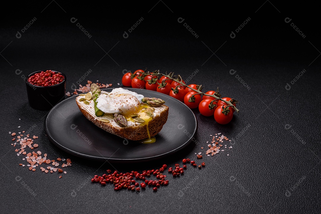 Delicioso café da manhã composto por ovos escalfados com torradas com cream cheese, aspargos, sal e especiarias em um fundo escuro de concreto