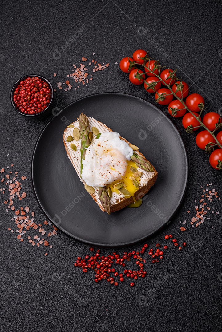 Delicioso café da manhã composto por ovos escalfados com torradas com cream cheese, aspargos, sal e especiarias em um fundo escuro de concreto