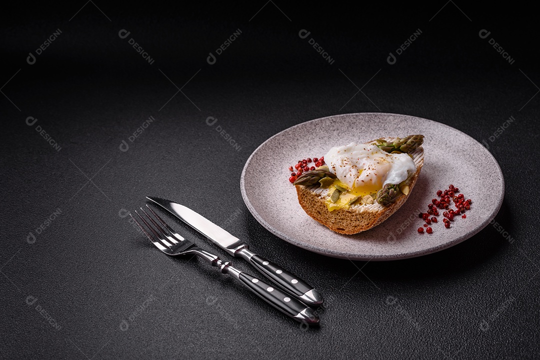 Delicioso café da manhã composto por ovos escalfados com torradas com cream cheese, aspargos, sal e especiarias em um fundo escuro de concreto