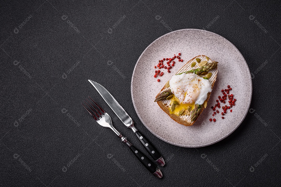 Delicioso café da manhã composto por ovos escalfados com torradas com cream cheese, aspargos, sal e especiarias em um fundo escuro de concreto